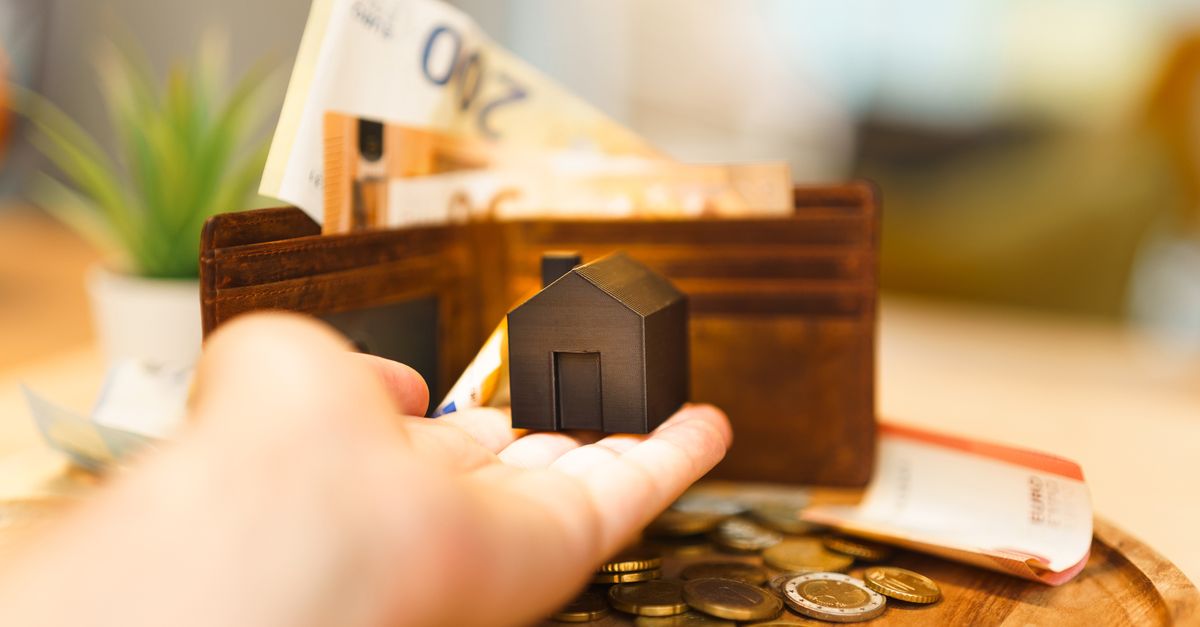 A hand holding a small house model with euro notes and coins nearby, illustrating real estate investment and finance.
